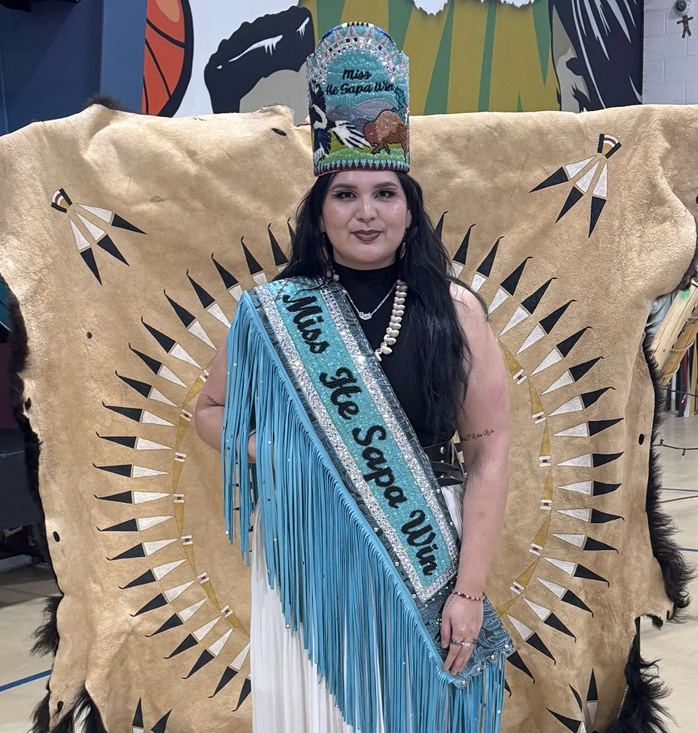 Young Native woman in formal dress in front of a decorated buffalo skin wearing sequined Lakota headpiece and sash that says "Miss He Sapa Win"