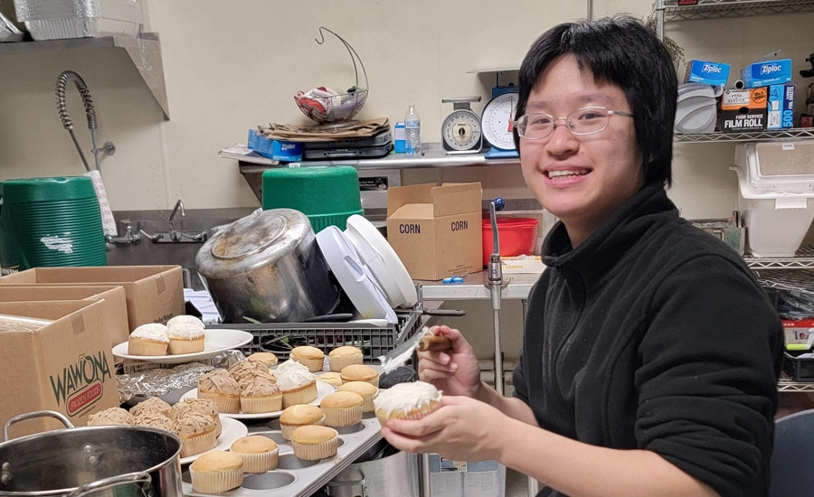 Young Asian person frosting cupcakes in the kitchen and smiling.