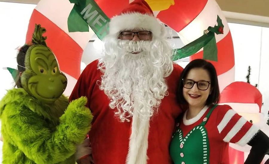 Adult woman dressed in holiday elf costume smiles next to people in Santa and Grinch costumes.