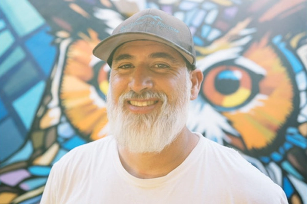 Smiling man with white beard and baseball cap in front of a colorful owl mural.