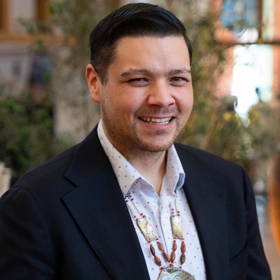 Smiling man with collared shirt and sport jacket and ceremonial necklace.