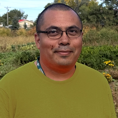 Smiling Native man with shaved head and olive colored tshirt with garden in background